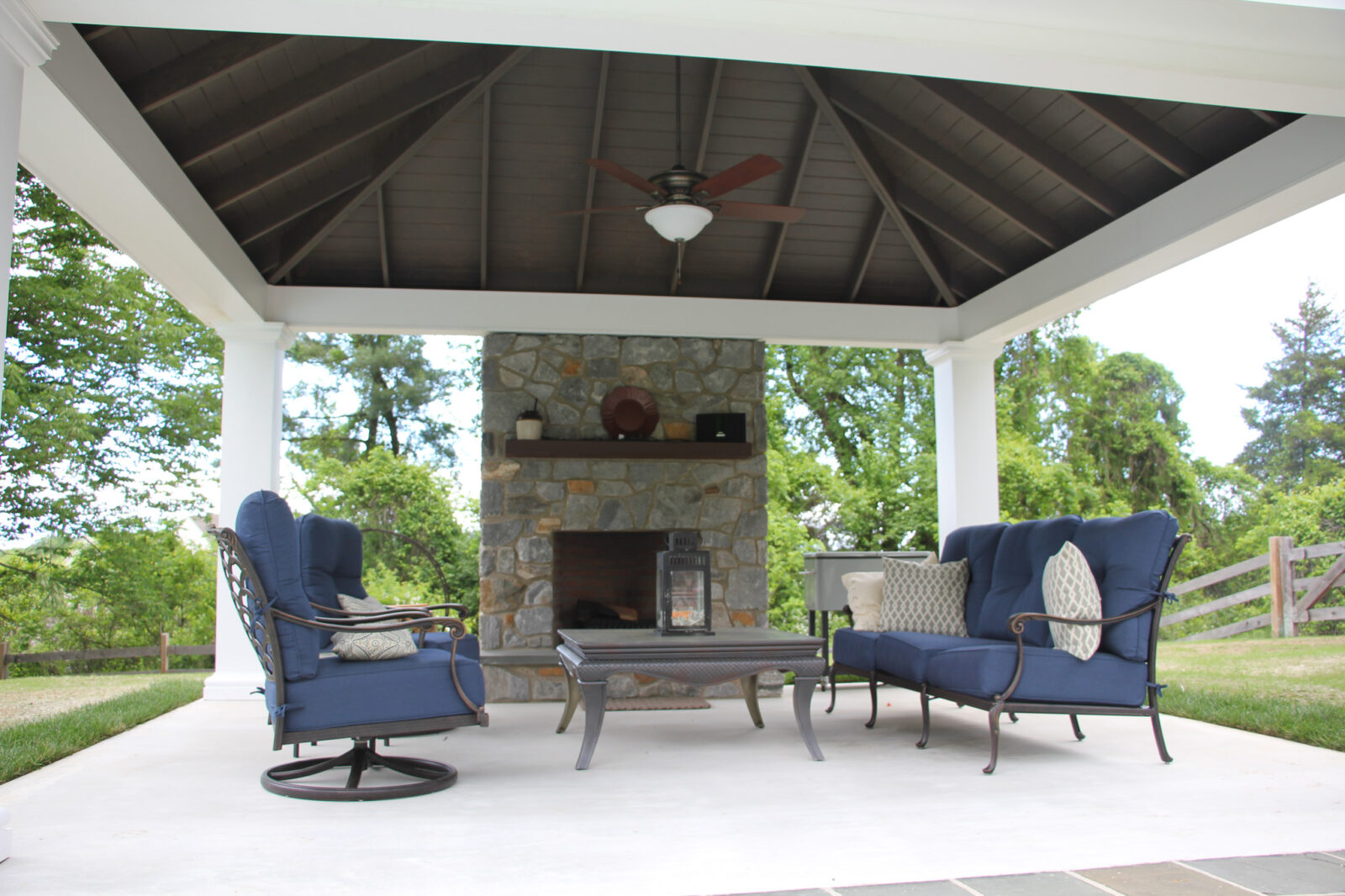 Lounge area in front of grey stone fireplace built into pavilion with ceiling fan and light